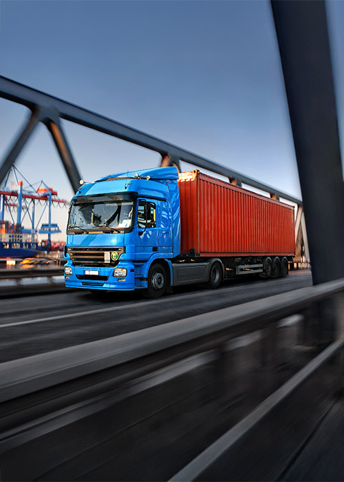 Lkw fährt über eine Brücke, im Hintergrund ist ein Containerschiff zu sehen